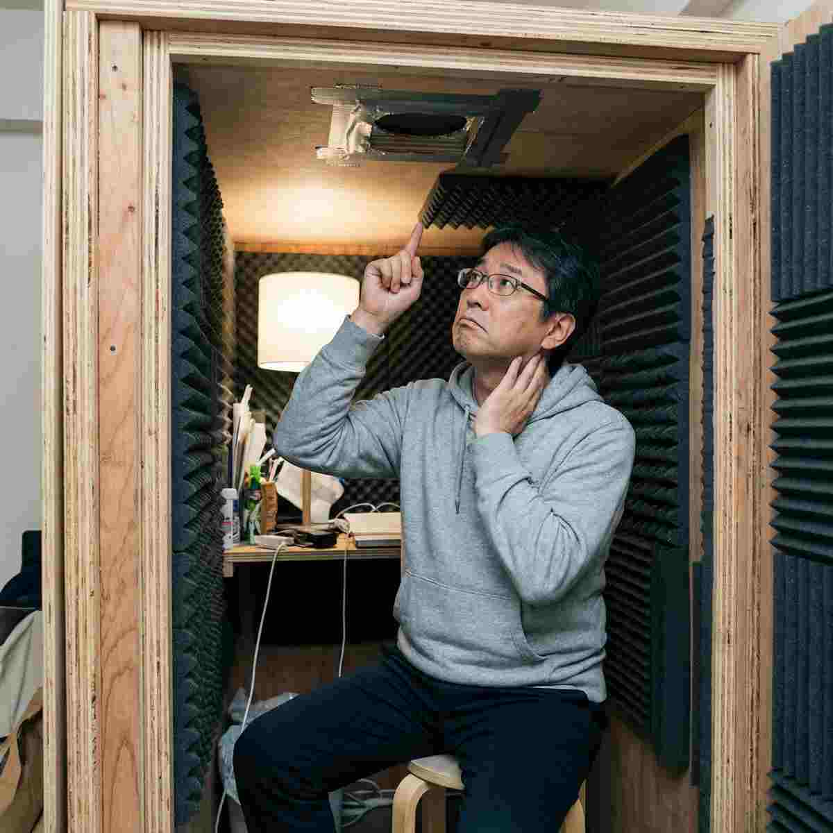 完成したDIY防音室の内部に座り、天井付近の換気口の隙間を指差して確認する男性